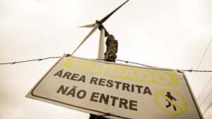 Placa de advertência vista de baixo para cima, inclinada contra um céu nublado. A placa está presa a um poste e traz os dizeres em letras maiúsculas: “CUIDADO – ÁREA RESTRITA – NÃO ENTRE”, acompanhados do ícone de uma pessoa com um traço diagonal cruzando, indicando proibição. Acima da placa, é possível ver parte de uma hélice de turbina eólica, sugerindo que a área é uma zona de geração de energia. Um pedaço de arame farpado atravessa horizontalmente o topo da imagem, reforçando a ideia de área protegida. A iluminação é suave e dourada.