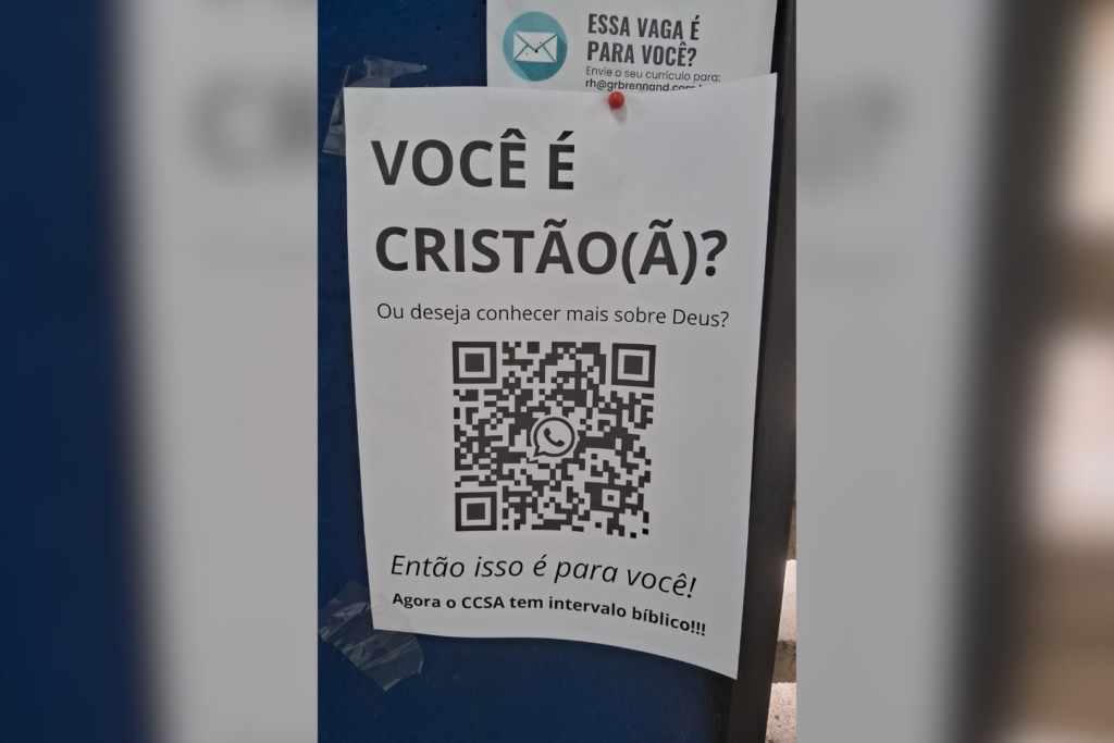 Esta imagem mostra um cartaz afixado em uma superfície azul. O cartaz contém um texto em português e um código QR. A mensagem principal convida cristãos ou pessoas interessadas em conhecer mais sobre Deus para participar de um intervalo bíblico no Centro de Ciências Sociais Aplicadas da UFPE. No cartaz, há um código QR com o logotipo do WhatsApp no centro. Acima do cartaz, parcialmente visível, há um outro papel que parece ser um anúncio de vaga de emprego. Ele convida interessados a enviarem currículos para um e-mail específico.