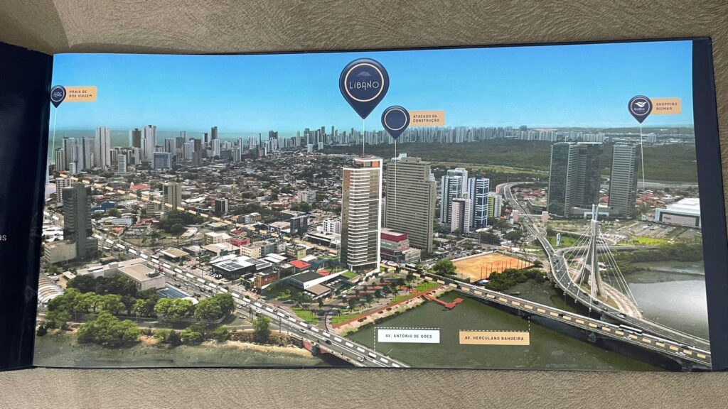Esta é uma reprodução de um material gráfico. Trata-se de uma imagem aérea de uma cidade com muitos prédios modernos. Ela destaca três locais importantes: a Praia de Boa Viagem à esquerda, um edifício chamado Líbano no centro, e o Shopping Riomar à direita. No centro, há um prédio alto identificado como Líbano. Abaixo dele, duas avenidas estão marcadas: Avenida Antônio de Goes e Avenida Herculano Bandeira. À direita, há uma ponte moderna conectando diferentes partes da cidade. A imagem transmite uma visão clara da infraestrutura urbana e destaca pontos de referência importantes, como a praia e o shopping. É uma representação detalhada da cidade, oferecendo um panorama interessante da organização dos espaços e do fluxo urbano.