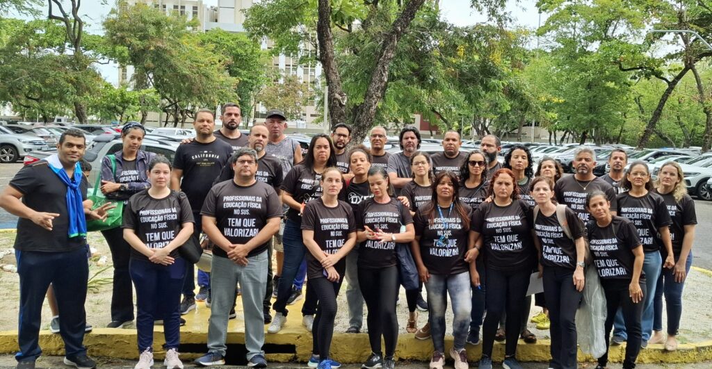 Cerca de 30 pessoas, homens e mulheres, estão reunidas em frente a um estacionamento, com expressão séria. Todos vestem camisas pretas com a frase: “Profissionais da Educação Física no SUS. Tem que valorizar!”. Algumas árvores e prédios aparecem ao fundo. O clima da imagem transmite união e reivindicação por respeito e valorização.