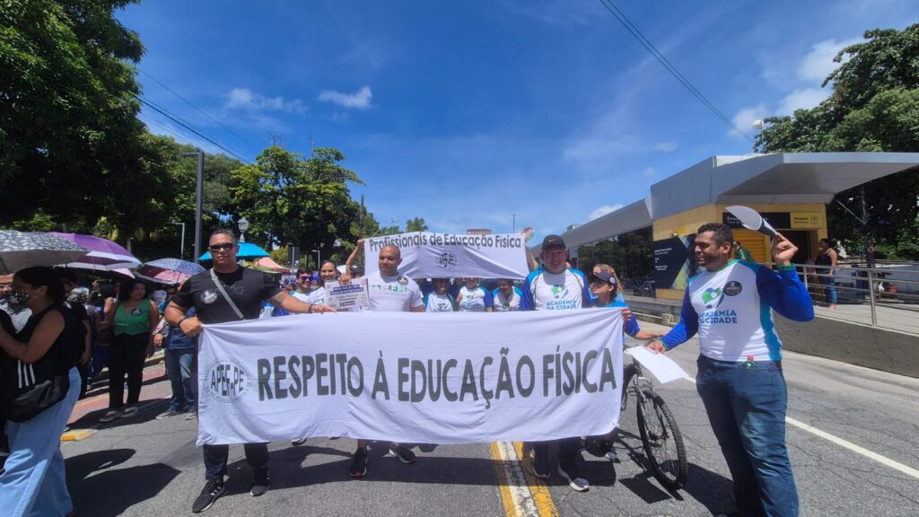 Um grupo de profissionais de educação física em uma manifestação ao ar livre, carregando uma faixa que diz “Respeito à Educação Física”. A maioria veste camisas brancas com azul e o logotipo do programa “Academia da Cidade”.