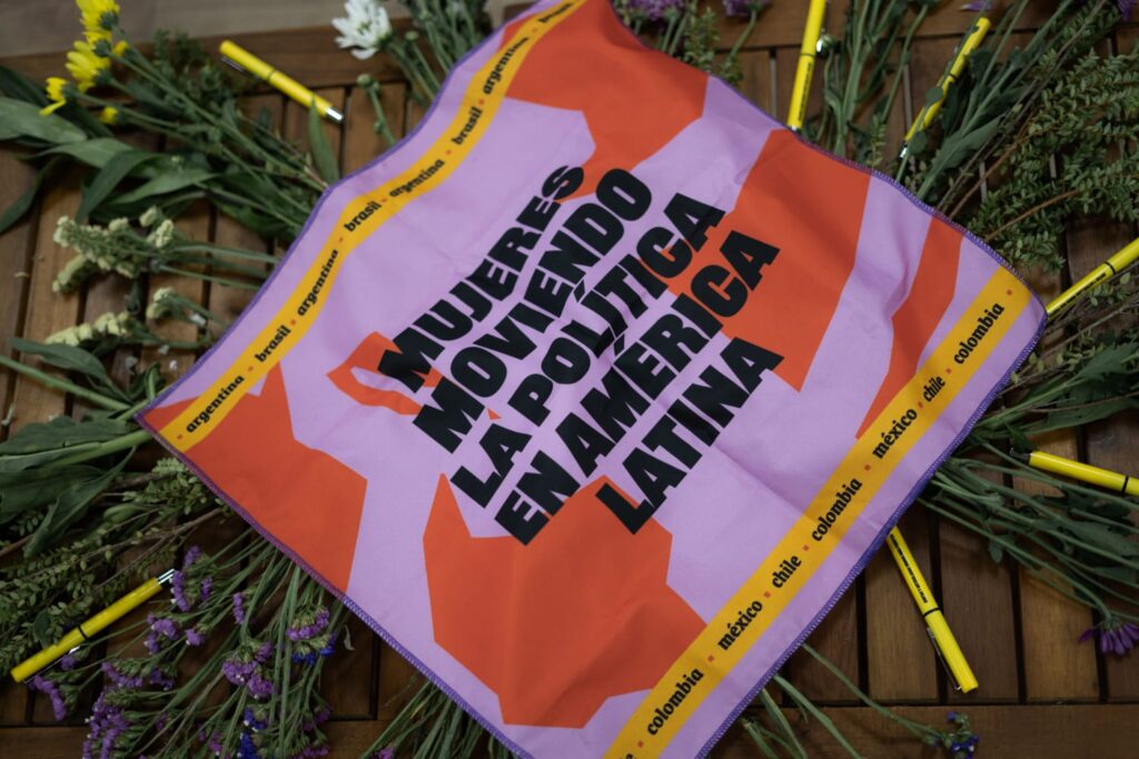A imagem mostra um lenço colorido sobre uma mesa de madeira, rodeado por flores e canetas amarelas. No centro do lenço, lê-se a frase: Mujeres moviendo la política en América Latina. O fundo é lilás com formas em laranja. Nas bordas, aparecem os nomes dos países: Argentina, Brasil, Colômbia, México e Chile. A composição transmite força e organização feminina na política latino-americana.