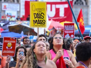 A imagem mostra uma manifestação com muitas pessoas reunidas na rua, segurando cartazes coloridos com mensagens. Um dos cartazes mais visíveis diz: Queremos ser pais e mães presentes, destacando a luta por melhores condições de trabalho e vida familiar. As pessoas estão atentas e engajadas, e há bandeiras e faixas ao fundo, incluindo uma grande com as letras CUT, que representa a Central Única dos Trabalhadores, uma organização sindical no Brasil. O clima é de mobilização social e reivindicação de direitos.