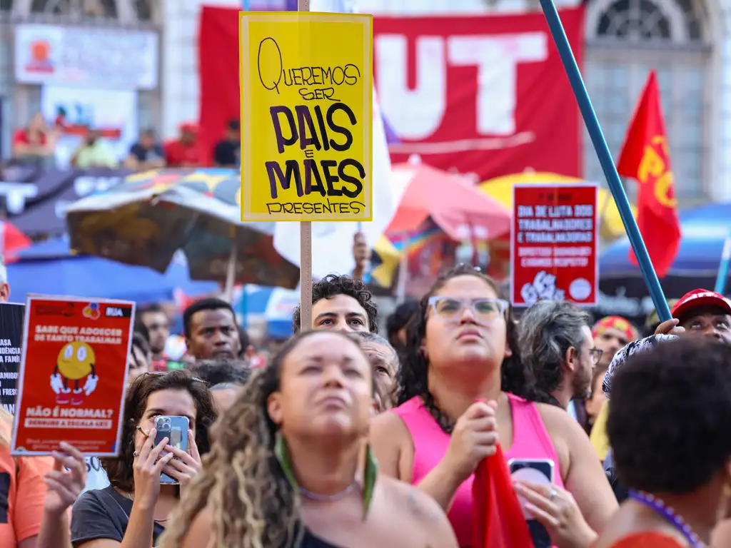 A imagem mostra uma manifestação com muitas pessoas reunidas na rua, segurando cartazes coloridos com mensagens. Um dos cartazes mais visíveis diz: Queremos ser pais e mães presentes, destacando a luta por melhores condições de trabalho e vida familiar. As pessoas estão atentas e engajadas, e há bandeiras e faixas ao fundo, incluindo uma grande com as letras CUT, que representa a Central Única dos Trabalhadores, uma organização sindical no Brasil. O clima é de mobilização social e reivindicação de direitos.