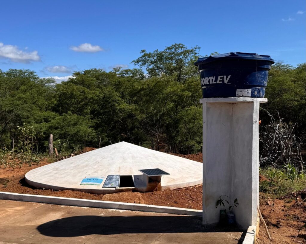 A fotografia mostra uma cisterna de captação de água da chuva em um ambiente rural, sob um céu azul com algumas nuvens esparsas. A cisterna, feita de concreto, tem formato circular e tampa cônica clara, levemente inclinada. Ao lado dela, sobre uma pequena base elevada de concreto, há uma caixa d’água azul da marca “Fortlev”. Um pequeno painel solar está instalado próximo à tampa da cisterna, indicando o uso de energia solar no sistema. A vegetação ao fundo é densa, com árvores típicas da caatinga. O solo ao redor é de terra avermelhada, característica de regiões semiáridas.