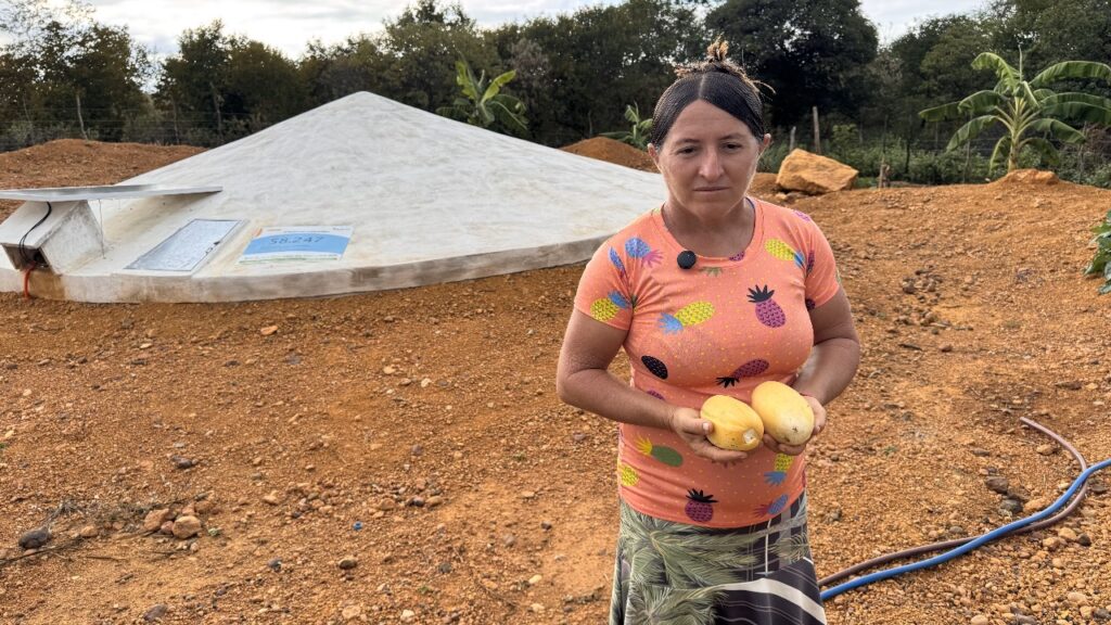 A fotografia mostra a agricultora Raimunda Juliana Coelho em um terreno de terra avermelhada, ao ar livre, durante o dia. Ela está em pé e olha para a câmera com expressão neutra. Raimunda veste uma blusa rosa estampada com abacaxis coloridos e uma saia com estampa de folhagens. Ela segura duas frutas amarelas, aparentemente mangas, nas mãos. Ao fundo, vê-se uma cisterna de concreto de formato circular com tampa inclinada, utilizada para captação de água da chuva — possivelmente parte de um projeto de cisterna com placas solares. Há vegetação esparsa e algumas bananeiras ao fundo, além de canos no chão à direita da imagem.