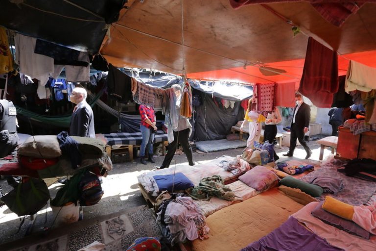 A imagem retrata o interior de um presídio em Pernambuco com diversas barracas protegidas por lonas e tecidos coloridos. Há homens caminhando pelo corredor e outros conversando. No chão, há itens variados, como roupas, cobertores e almofadas, A iluminação mistura sombras e pontos de luz natural que atravessam as aberturas das lonas, criando uma atmosfera semelhante a de um mercado ou feira livre.