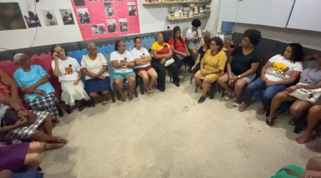 A imagem mostra um grupo de mulheres sentadas em círculo em uma sala simples e comunitária. Elas estão acomodadas em cadeiras de plástico, aparentemente participando de uma reunião ou discussão. No fundo, há uma parede com várias fotos e cartazes afixados, alguns em um painel vermelho. Também é possível ver uma prateleira contendo livros e alguns armários. O chão é de concreto, reforçando a sensação de um ambiente funcional e acessível. Parece ser um espaço usado para encontros e troca de ideias.