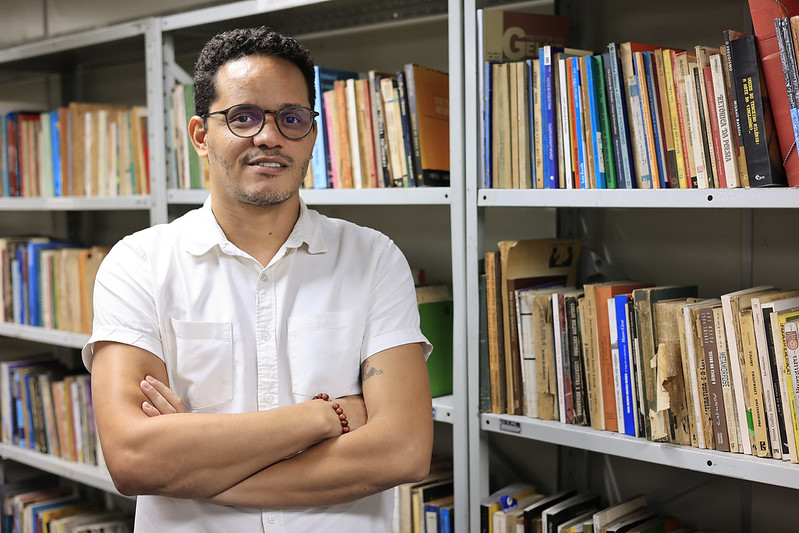 O professor Cleber Ataíde está de pé em frente a estantes metálicas repletas de livros, em um ambiente que lembra um arquivo ou biblioteca universitária. Ele olha para a câmera com um leve sorriso, transmitindo serenidade e segurança. Cleber tem pele parda, cabelo curto e cacheado, e usa barba rala. Está com óculos de armação escura, redonda e translúcida. Ele veste uma camisa social branca de manga curta e está com os braços cruzados à frente do corpo. No pulso esquerdo, usa uma pulseira de contas marrons. Do lado interno do braço direito, aparece parte de uma tatuagem com linhas pretas. A iluminação é clara, destacando os livros ao fundo, que variam entre obras antigas e mais recentes, com capas de cores diversas.