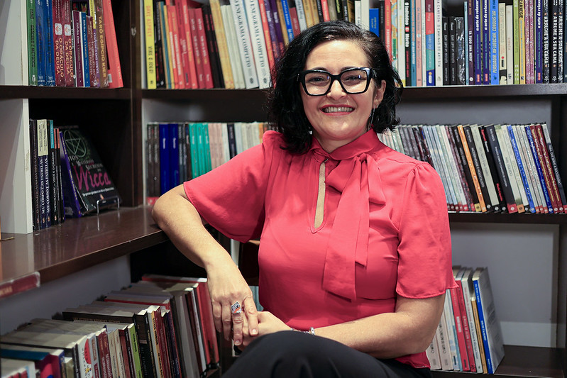 A professora Siane Gois está sentada em um ambiente que parece ser uma biblioteca ou escritório, cercada por estantes repletas de livros coloridos. Ela está sorrindo e olhando diretamente para a câmera, com uma expressão acolhedora e confiante. Siane tem pele clara, cabelo preto liso e na altura dos ombros, usado solto e penteado de lado. Usa óculos grandes de aro preto. Ela veste uma blusa rosa-choque com um laço no pescoço e uma abertura discreta na altura do colo. Está com as pernas cruzadas e as mãos apoiadas sobre o joelho, mostrando anéis prateados com pedras. O ambiente é bem iluminado, com luz suave que destaca os detalhes dos livros ao fundo, organizados por cor e tamanho.