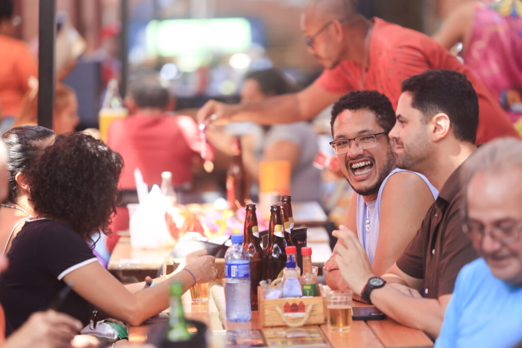 A foto mostra um grupo de pessoas reunidas em um ambiente descontraído, provavelmente um bar ou restaurante ao ar livre, durante o dia. Elas estão sentadas em mesas de madeira compridas, próximas umas das outras, interagindo animadamente. No centro da imagem, dois homens estão em destaque: um deles, com barba rala, óculos e aparelho nos dentes, está sorrindo amplamente e olhando diretamente para a câmera. O outro, ao seu lado direito, ouve atentamente e sorri levemente. Na mesa há várias garrafas de cerveja, uma garrafa de água, copos com bebidas e um recipiente com condimentos (molhos e temperos). Ao fundo, outras pessoas também conversam e um garçom de camiseta vermelha está servindo algo em um copo.
