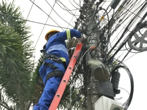 Na imagem, vemos um trabalhador vestido com um uniforme azul e capacete amarelo subindo uma escada vermelha encostada em um poste de concreto. Ele está realizando manutenção em uma fiação elétrica bastante desorganizada e caótica. O trabalhador está preso com um cinto de segurança, e há muitos cabos pretos emaranhados ao redor do poste, estendendo-se em várias direções. O cenário parece urbano, com folhas de palmeiras visíveis ao fundo, sugerindo que o local é em uma região tropical ou subtropical. A quantidade de fios parece excessiva e mal organizada, o que pode indicar problemas de infraestrutura elétrica ou de telecomunicação. O clima parece nublado ou o céu está claro, mas sem sol visível.