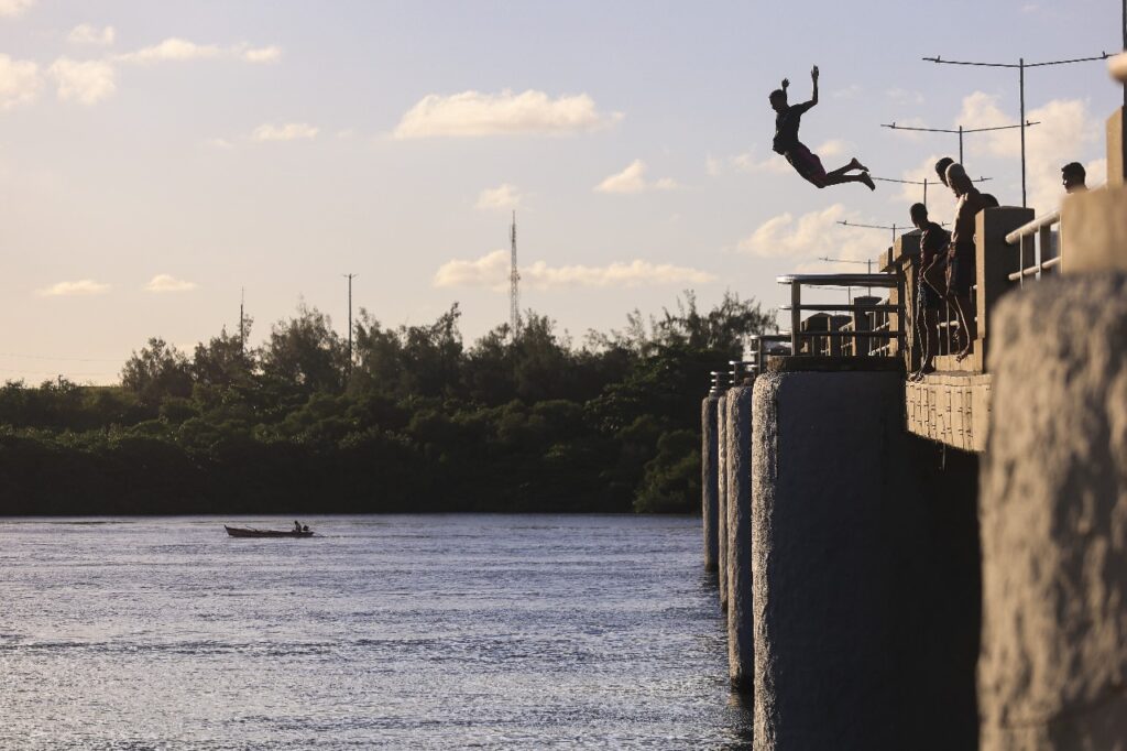 A foto mostra um grupo de jovens sobre uma ponte, durante o entardecer, à beira de um rio ou braço de mar. Um dos meninos está no ar, em pleno salto, com o corpo inclinado para trás e os braços abertos, prestes a cair na água. O céu está claro, com algumas nuvens e luz dourada típica do fim da tarde. Ao fundo, vê-se vegetação densa na margem oposta e um pequeno barco com duas pessoas navegando. A cena transmite leveza, liberdade e espírito de aventura, com destaque para o contraste entre as sombras da ponte e a claridade do céu.