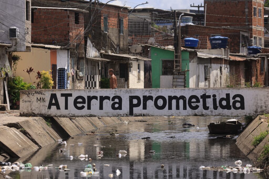 O ChatGPT disse: A foto mostra uma comunidade periférica com casas simples, muitas delas construídas com tijolos expostos e materiais improvisados. No centro da imagem, atravessando um canal poluído e repleto de lixo flutuante, há uma mureta branca com a frase escrita em letras grandes e pretas: “A Terra Prometida”. Atrás da mureta, um homem sem camisa caminha pela calçada. As construções ao fundo têm aspecto precário, com fachadas desgastadas, fios elétricos expostos, caixas d’água azuis nos telhados e grades nas janelas.