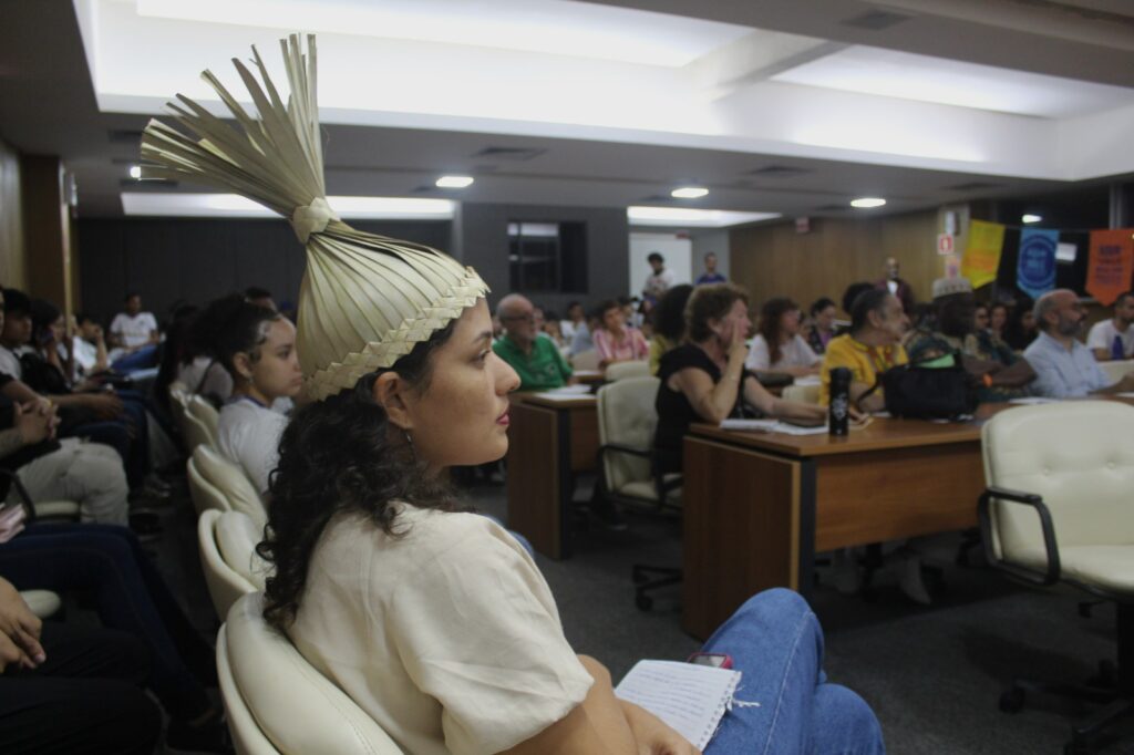Em um auditório da Fundação Joaquim Nabuco (Fundaj), no Recife, uma mulher aparece em primeiro plano, sentada e atenta ao que acontece no evento. Ela tem pele clara, cabelos cacheados escuros e usa um cocar tradicional feito de palha. Está com uma caderneta no colo e um celular ao lado. Ao fundo, várias pessoas de diferentes idades e origens também assistem à programação, sentadas em cadeiras claras. Algumas fazem anotações ou observam atentamente. O ambiente é iluminado por luzes brancas de teto e tem mesas de madeira.