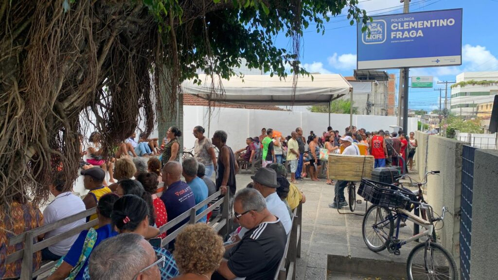 Esta foto mostra uma cena ao ar livre, em frente à Policlínica Clementino Fraga, localizada na cidade do Recife, como indica a placa azul no canto superior direito da imagem. A cena está bastante movimentada, com uma grande quantidade de pessoas reunidas. No lado esquerdo da imagem, há muitas pessoas sentadas em bancos de madeira, aparentemente esperando atendimento. São em sua maioria idosos, vestidos de forma simples, e a maioria está à sombra de uma grande árvore com raízes expostas, que cobre parcialmente a área. No centro e à direita da imagem, há um grupo maior de pessoas em pé, algumas parecendo estar em fila ou aglomeradas. Algumas estão interagindo, outras parecem estar esperando. Ao fundo, há uma estrutura coberta que abriga mais pessoas sentadas.