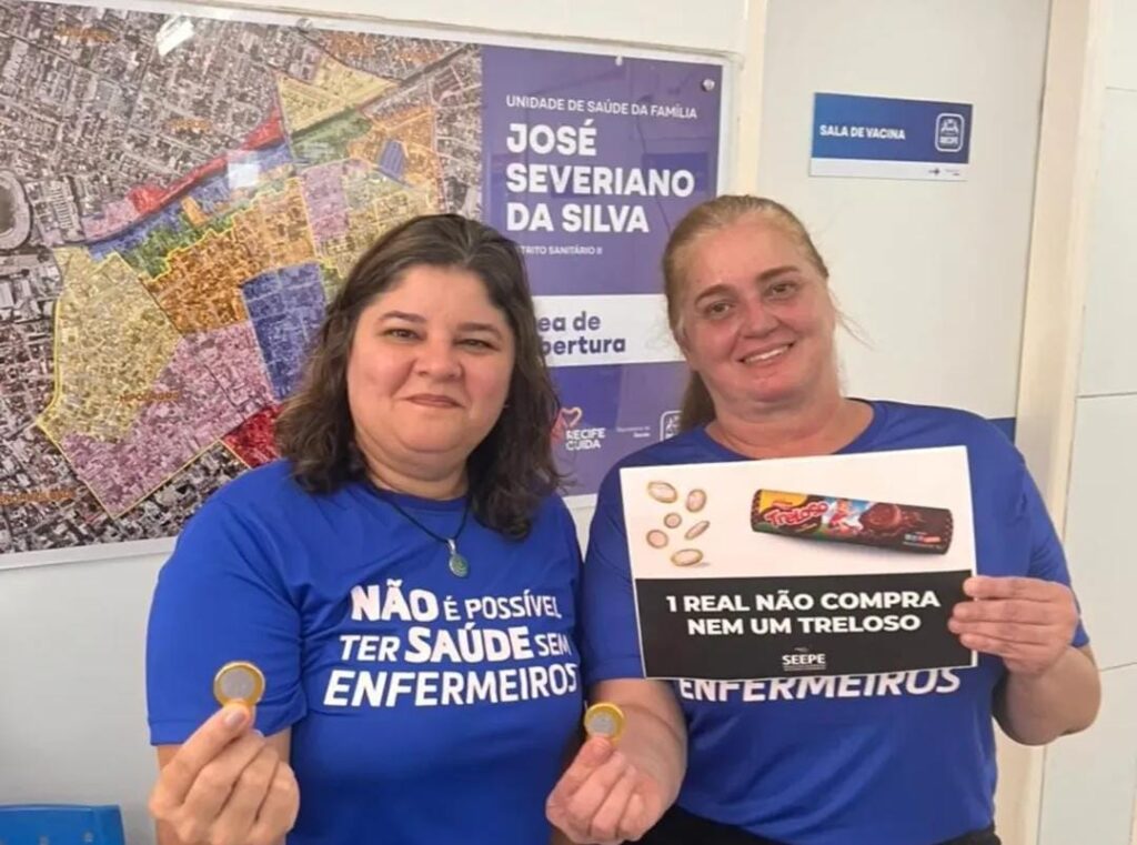 A imagem mostra duas mulheres de meia idade usando camisetas azuis onde se lê a frase não é possível ter saúde sem enfermeiros em letras brancas. A mulher à direita da imagem segura um pequeno cartaz com uma foto de um pacote de biscoito da narca treloso e a frase 1 real não compra nem um treloso. As duas foram fotografadas em um ambiente interno onde se vê um banner indicando a unidade de saúde da família José Severiano da Silva.