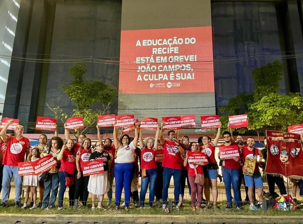 Foto noturna de um grande grupo de pessoas usando camisetas vermelhas com estampas variadas levantando pequenos cartazes vermelhos com a palavra greve em letras brancas acima das cabeças. As pessoas estão em frente a uma grande parede de concreto cinza onde está projetada a frase educação do Recife está em greve! João Campos, a culpa é sua.