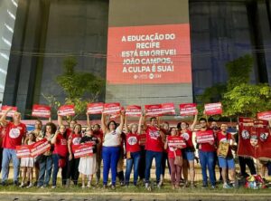 Foto noturna de um grande grupo de pessoas usando camisetas vermelhas com estampas variadas levantando pequenos cartazes vermelhos com a palavra greve em letras brancas acima das cabeças. As pessoas estão em frente a uma grande parede de concreto cinza onde está projetada a frase educação do Recife está em greve! João Campos, a culpa é sua.
