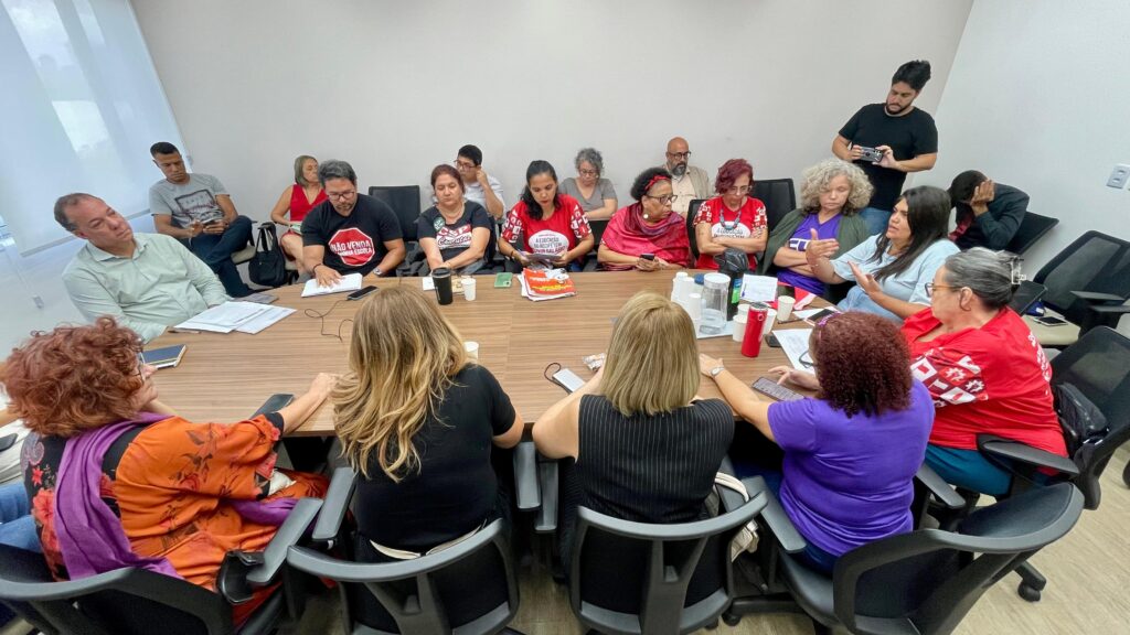 A foto mostra um grupo de cerca de 18 pessoas reunidas em uma sala de reunião, todas concentradas em uma grande mesa. Muitas delas estão vestindo camisetas vermelhas com textos e logotipos, sugerindo que pertencem a uma mesma organização ou evento. Algumas pessoas estão sentadas, outras em pé, e todas parecem envolvidas em alguma atividade, seja conversando, tomando notas ou verificando seus celulares. A mesa está cheia de objetos, como papéis, copos e garrafas de água, indicando que a reunião envolve discussões e planejamento. A sala é bem iluminada e há uma janela grande ao lado esquerdo, deixando entrar bastante luz natural.