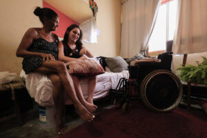 Nesta foto, vemos duas mulheres sentadas em uma cama simples dentro de um quarto iluminado por luz natural vinda de uma janela com cortinas brancas. A mulher mais velha, à esquerda, é Ester da Silva; ela está usando um vestido escuro de alças e está descalça. A mais jovem, à direita, é sua filha Evellyn Vitória , que segura uma almofada no colo e sorri com carinho para a mãe. Há um clima de intimidade e afeto entre elas, evidenciado pelo sorriso e toque físico. No canto direito da imagem, há uma cadeira de rodas, usada pela jovem, que tem mobilidade reduzida. O ambiente é simples, mas acolhedor, com plantas, almofadas e uma atmosfera caseira. A imagem transmite uma sensação de apoio mútuo, cuidado familiar e afeto, mesmo em meio a condições modestas de moradia.