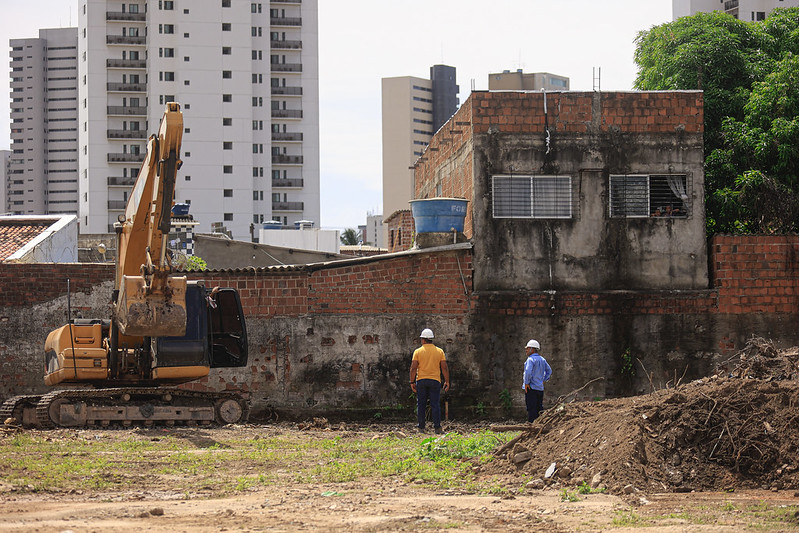 A imagem mostra um local de construção. À esquerda, há uma escavadeira amarela, parada em um terreno de terra, indicando que a obra está em andamento. Dois trabalhadores estão de pé no meio do cenário, usando capacetes de segurança. Um deles veste uma camisa amarela e calças escuras, enquanto o outro usa uma camisa azul e calças escuras. Eles parecem observar uma parede de tijolos. Atrás da parede de tijolos, há uma casa de alvenaria com duas janelas protegidas por grades. No telhado dessa casa, há uma caixa d'água azul. O fundo da imagem revela vários prédios altos, sugerindo que essa obra acontece em uma área urbana.