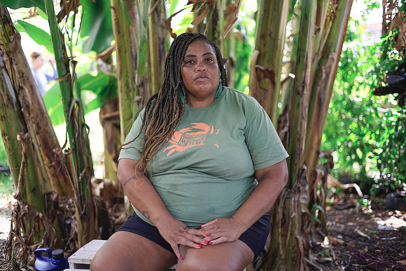 Foto de Sarah Marques, mulher negra, com tranças no cabelo castanho, usando camiseta verde e bermuda escura. Ela está sentada, com as mãos pousadas sobre o colo, em um ambiente ao ar livre na frente de algumas babaneiras, tendo ao fundo vegetação que está desfocada na imagem.