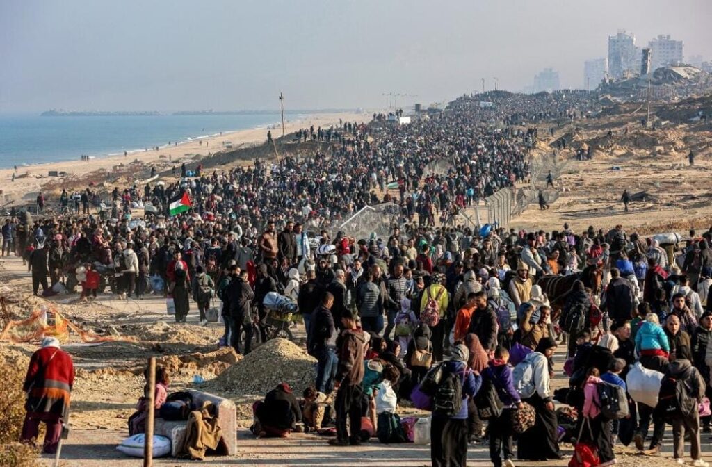 A imagem mostra uma grande multidão de pessoas reunidas em uma área aberta próxima à praia. Trata-se de milhares de palestinos retornando para suas casas em Gaza. Uma cerca separa o grupo em duas partes, com muitas pessoas de um lado e algumas do outro. Ao fundo, vê-se a praia e o mar, além de alguns prédios ao longe. Muitas pessoas carregam mochilas e sacolas, e algumas estão sentadas ou deitadas no chão. Entre a multidão, há uma bandeira palestina visível.