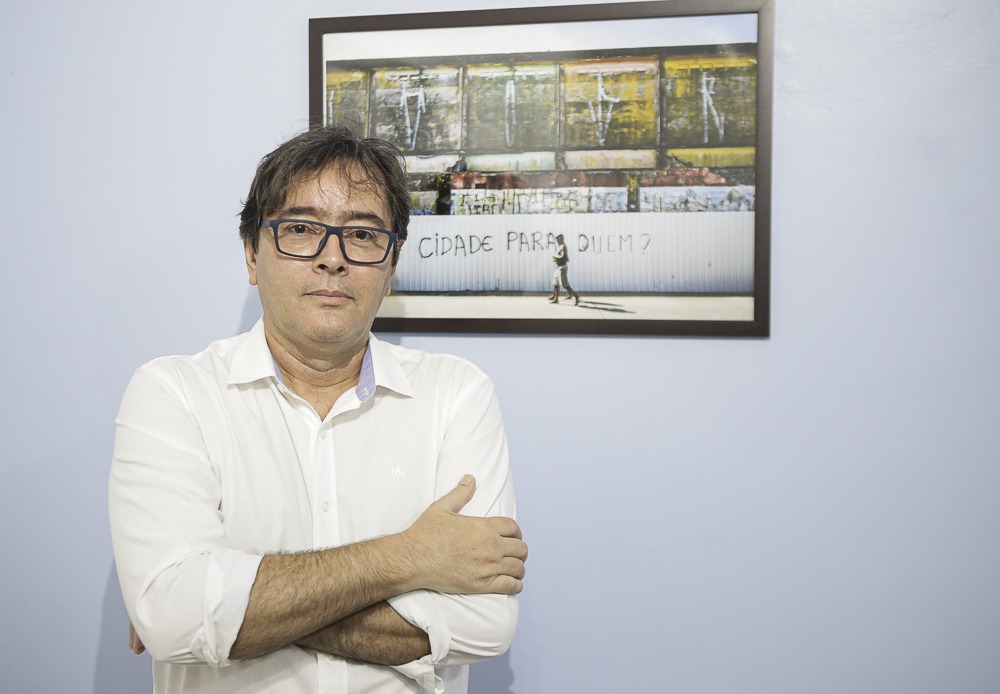 Foto de Fabiano Rocha Diniz, homem de pele clara, cabelo castanho escuro e óculos de armação preta. Ele está vestindo uma camisa social branca e está com os braços cruzados, apoiando o braço esquerdo com a mão direita. Seu rosto tem uma expressão séria e calma. Ele está em pé em frente a uma parede de cor azul clara. Atrás dele, preso à parede, há um quadro com uma fotografia colorida. A imagem dentro do quadro mostra um muro branco com a frase pichada: CIDADE PARA QUEM?, e há uma pessoa andando na calçada em frente a esse muro. Acima do muro, há uma estrutura com contêineres empilhados, enferrujados e desgastados