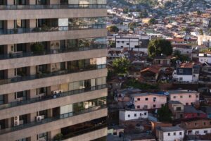 A imagem mostra um forte contraste urbano entre dois mundos vizinhos. À esquerda, ocupa quase metade da foto um prédio moderno de apartamentos, alto, com fachada cinza, varandas com grades de vidro e ar-condicionado visível em várias unidades. As janelas são grandes e fechadas, transmitindo um ar de ordem e privacidade. À direita da imagem, estende-se uma comunidade com casas simples e irregulares, construídas com tijolos expostos, telhados de diferentes alturas e cores. As casas estão muito próximas umas das outras, formando um cenário denso e popular. Há roupas penduradas, árvores esparsas e vegetação entre as construções. Ao fundo, é possível ver a paisagem urbana continuar por sobre morros e colinas, revelando a extensão da cidade.