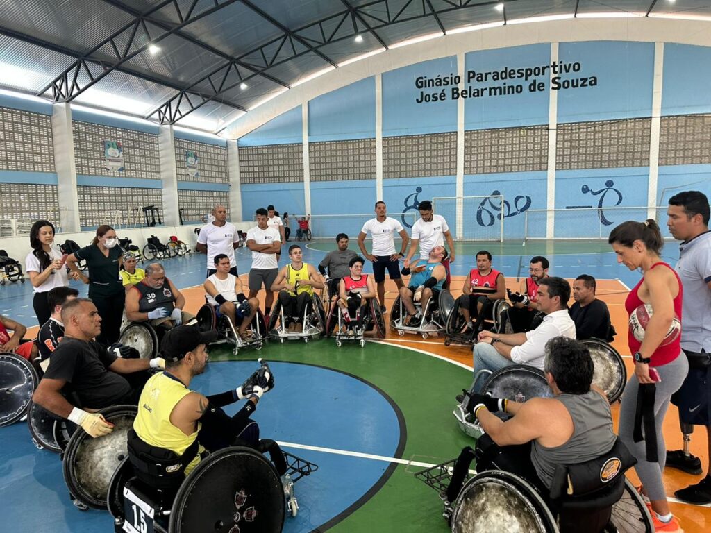 A imagem mostra um grupo de pessoas em cadeiras de rodas reunidas em círculo dentro de um ginásio esportivo, que tem o nome Ginásio Paradesportivo José Belarmino de Souza escrito na parede ao fundo. Os participantes parecem envolvidos em uma atividade ou discussão, com alguns vestindo uniformes esportivos e outros roupas casuais. Ao redor do círculo, há pessoas em pé que podem ser treinadores ou assistentes. O ambiente é iluminado, e o piso da quadra tem marcações coloridas, indicando seu uso para diferentes esportes.