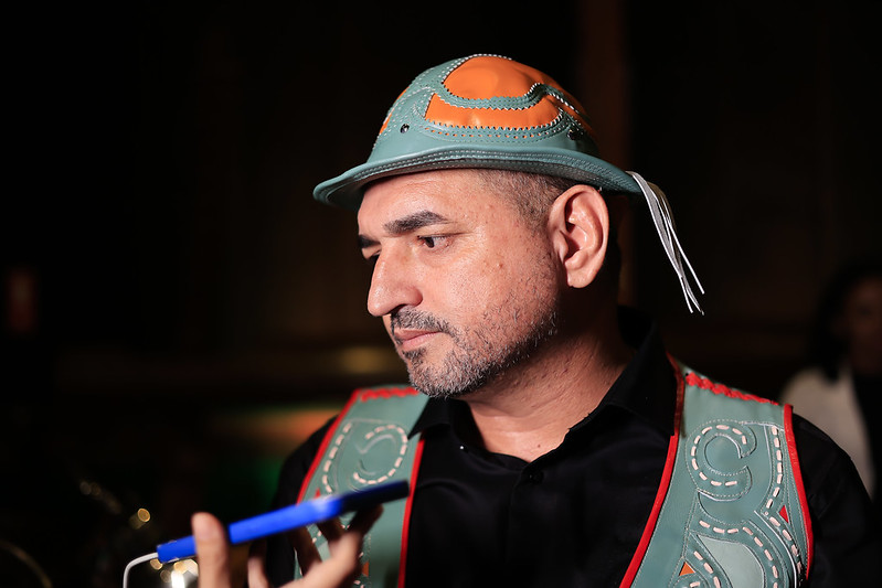 A foto mostra Aleudo Benedito, prefeito de Serrita (PE), em destaque, usando traje típico de vaqueiro nordestino. Ele veste um colete de couro verde e laranja com bordados e usa um chapéu de couro combinando. Está em ambiente interno com iluminação quente, que destaca seu rosto e roupas. Aleudo olha sério para a esquerda, enquanto responde a uma entrevista. Um celular azul aparece próximo a ele, com uma mão segurando o aparelho celular. O fundo está escuro e desfocado, mantendo o foco totalmente sobre ele.
