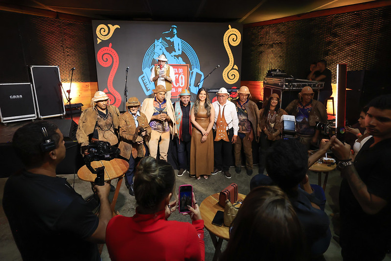 A foto mostra um grupo de pessoas posando para uma foto em um evento culturalno Nordeste brasileiro. Ao fundo, há um painel com elementos gráficos coloridos e o nome do evento: Festa do Jacó, junto com a imagem estilizada de um vaqueiro montado a cavalo, em azul. O cenário está iluminado com luzes quentes e acolhedoras. No centro da imagem, há 10 pessoas lado a lado, sorrindo e olhando para as câmeras. Muitas delas estão vestidas com roupas típicas de vaqueiro nordestino: chapéus de couro, gibões (espécie de casaco de couro), botas e roupas de tom terroso. Entre elas, há também mulheres com vestidos e trajes que combinam com o estilo regional, misturando tradição e modernidade. Alguns participantes seguram microfones, indicando que podem ser artistas ou representantes do evento. Na frente, várias pessoas estão tirando fotos e filmando o grupo com celulares e câmeras profissionais.