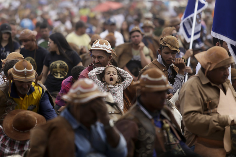 A foto mostra uma multidão participando de um evento tradicional no sertão, com muitos homens vestidos como vaqueiros nordestinos — usando chapéus e roupas de couro. Em destaque, um homem carrega uma menina no colo; ela está bocejando com os braços levantados sobre a cabeça. O clima é de celebração e devoção, sugerindo um momento de fé típico da missa do vaqueiro.