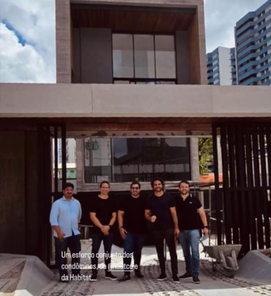 A foto mostra cinco homens posando sorridentes em frente à entrada de um edifício moderno recém-construído. Eles estão lado a lado, vestidos de forma casual, com camisas escuras e calças jeans, exceto um que usa camisa azul clara. Atrás deles, o prédio tem fachada de vidro e concreto com linhas retas e aparência sofisticada. Duas carriolas de obra ainda estão no local, sugerindo que a construção foi recentemente finalizada ou está nos ajustes finais. Ao fundo, vê-se parte do céu com nuvens e edifícios altos residenciais. No canto inferior esquerdo da imagem, há uma legenda escrita: “Um esforço conjunto dos condôminos, da @hautcce e da Habitat.”