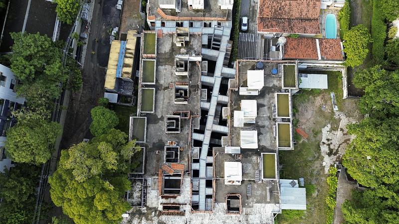 A imagem mostra uma vista aérea de um prédio em construção. A construção ocupa a maior parte do centro da imagem e tem um formato retangular com um corredor estreito e sinuoso no meio, criando uma aparência quase labiríntica. A estrutura ainda está inacabada — é possível ver os blocos de concreto, as paredes sem acabamento e áreas abertas onde os andares ainda estão sendo erguidos. Ao redor do prédio, há bastante vegetação e árvores, especialmente do lado esquerdo e na parte inferior da imagem. À direita, há casas térreas com telhados de telha vermelha, sendo que uma delas tem uma piscina retangular de cor azul no quintal. Há também uma área de terra batida, com grama esparsa, como se fosse um quintal ou lote vazio. A cena mostra claramente o contraste entre a construção urbana e o ambiente residencial com natureza ao redor, evidenciando um espaço em transformação.
