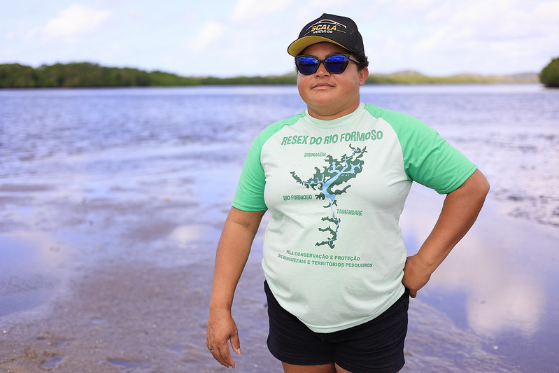 A foto mostra Cícera Estevão Batista em pé, em uma área de manguezal próxima à água, com o fundo formado por rio, céu azul e vegetação ao longe. Ela veste uma camiseta verde clara com mangas verde-escuras, que traz o mapa da Reserva Extrativista (Resex) do Rio Formoso e os nomes das localidades Rio Formoso e Tamandaré, além da frase: “Pela conservação e proteção das marisqueiras e territórios pesqueiros.” Cícera usa boné preto com detalhes amarelos, óculos escuros e bermuda preta. Ela tem expressão firme e postura confiante, com uma das mãos na cintura e a outra ao lado do corpo.