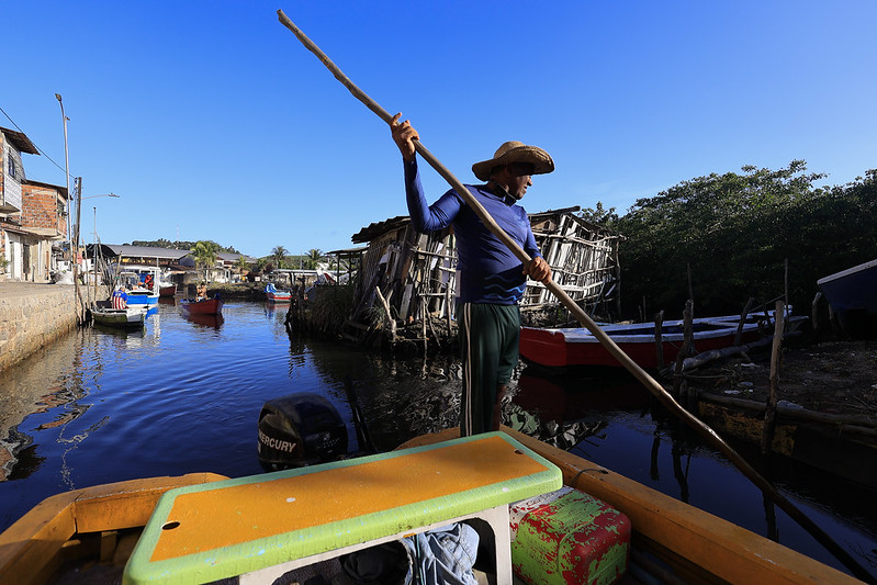 A foto mostra um homem em pé na parte de trás de um pequeno barco de madeira, conduzindo-o com um longo remo. Ele veste camisa azul escura de manga comprida, calça verde e chapéu de palha de abas largas. A embarcação está em um canal estreito, cercado por casas simples à esquerda e vegetação à direita. À margem do canal, há outras embarcações atracadas, algumas de madeira colorida. Uma construção rústica feita de tábuas de madeira aparece em destaque ao fundo, parcialmente inclinada. O céu está limpo, em um tom azul intenso, e a luz do sol ilumina fortemente a cena.