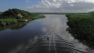 A foto mostra uma vista aérea de um largo rio cercado por uma vegetação densa de manguezais e morros cobertos de árvores. No centro da imagem, dois pequenos barcos navegam lado a lado, deixando rastros simétricos na água escura e espelhada. O céu está parcialmente nublado, com nuvens brancas e cinzentas refletidas na superfície do rio, criando uma atmosfera serena. À esquerda, há um morro mais alto com vegetação exuberante e uma encosta parcialmente exposta. Ao fundo, a paisagem se estende até o horizonte, com áreas verdes, planícies e céu aberto.