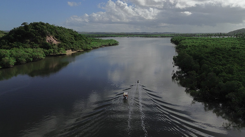 A foto mostra uma vista aérea de um largo rio cercado por uma vegetação densa de manguezais e morros cobertos de árvores. No centro da imagem, dois pequenos barcos navegam lado a lado, deixando rastros simétricos na água escura e espelhada. O céu está parcialmente nublado, com nuvens brancas e cinzentas refletidas na superfície do rio, criando uma atmosfera serena. À esquerda, há um morro mais alto com vegetação exuberante e uma encosta parcialmente exposta. Ao fundo, a paisagem se estende até o horizonte, com áreas verdes, planícies e céu aberto.