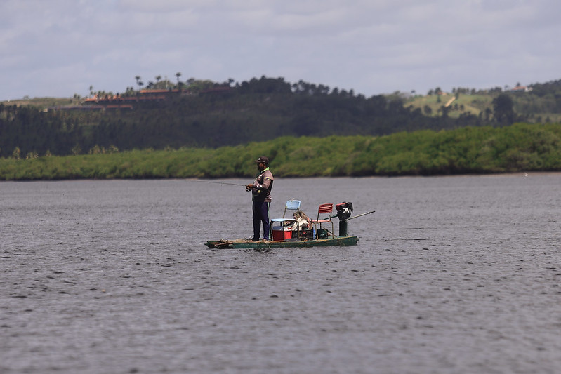 A foto mostra um pescador negro em pé sobre uma pequena balsa ou jangada de madeira no meio de um rio. Ele veste colete, boné e segura uma vara de pescar, concentrado na atividade. A embarcação é simples e improvisada, equipada com duas cadeiras coloridas, um motor na parte de trás e alguns baldes e utensílios de pesca espalhados ao redor. O rio está calmo, e ao fundo há uma vegetação verde densa, típica de áreas de mangue, além de morros cobertos por mata e algumas construções distantes. O céu está nublado, suavizando a luz da cena.