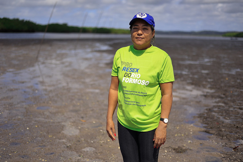 A foto mostra Arlene Maria da Costa em pé em uma área de manguezal durante a maré baixa, com o solo coberto por lama escura e alguns reflexos d'água ao fundo. Ela usa uma camiseta verde-limão com inscrições azuis e brancas que promovem a “RESEX do Rio Formoso” e mencionam eventos internacionais voltados à pesca artesanal e à preservação dos manguezais. Arlene veste ainda um boné azul com símbolo branco, calça escura e relógio no pulso. Seu rosto tem expressão serena e determinada, e o fundo da imagem mostra vegetação de mangue e céu parcialmente nublado.