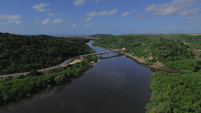 A imagem aérea mostra um rio largo e sinuoso cortando uma paisagem coberta por vegetação verde e densa. No centro da cena, há uma ponte de concreto que cruza o rio, ligando duas margens formadas por morros cobertos de árvores. À esquerda da ponte, há uma estrada que acompanha a margem do rio e algumas construções pequenas próximas à água. Também é possível ver embarcações atracadas próximo à ponte, sugerindo uma área de parada ou porto local. Ao fundo, o horizonte revela uma área urbana com construções baixas, contrastando com o verde predominante. O céu está azul, com nuvens brancas dispersas.