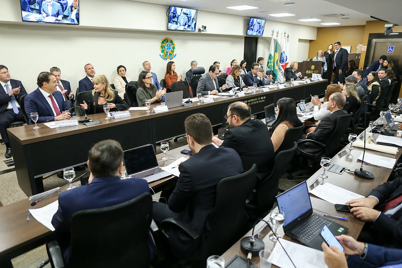 A foto mostra uma reunião formal da Ordem dos Advogados do Brasil (OAB), realizada em uma ampla sala com mesa em formato de “U”. Várias pessoas, a maioria em trajes formais como ternos e vestidos sociais, estão sentadas ao redor da mesa. Na parte central da imagem, há uma fileira de autoridades ou conselheiros da OAB, com microfones à frente, alguns em postura de fala, outros ouvindo ou aplaudindo. Atrás deles, na parede, vê-se o brasão da República Federativa do Brasil, televisores ligados e várias bandeiras oficiais, incluindo a do Brasil. O ambiente é bem iluminado e passa uma atmosfera institucional e séria