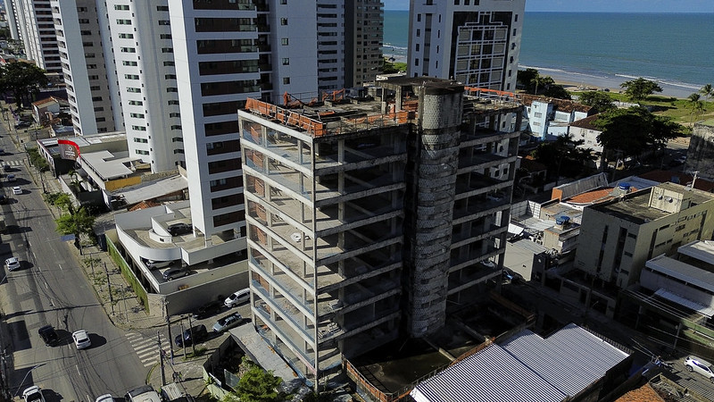 A foto mostra um prédio alto e inacabado, visto de cima por um drone, localizado em uma área urbana próxima à praia. A construção tem cerca de dez andares, com a estrutura de concreto exposta, sem janelas ou acabamentos, e um grande cilindro vertical de concreto ao centro, provavelmente o poço do elevador. Há uma tela laranja de proteção ao redor do topo da obra. Ao redor do prédio, há outros edifícios altos, modernos e finalizados, formando uma paisagem urbana densa. À esquerda, uma avenida movimentada com carros e calçadas largas acompanha a linha de prédios. Ao fundo, vê-se o mar azul e uma faixa de areia clara com algumas árvores, indicando proximidade com a orla. O contraste entre o prédio em obras e os arranha-céus modernos evidencia a transição ou abandono daquela estrutura.