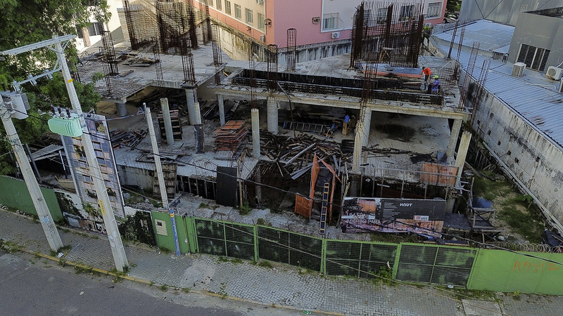 A imagem mostra uma construção inacabada, com estrutura de concreto e vergalhões expostos, indicando que a obra foi interrompida antes de avançar. Há muito entulho, madeiras e ferros espalhados pelo canteiro. Dois trabalhadores com capacetes aparecem na parte superior da laje. O local é cercado por um muro verde com grades e está localizado em uma calçada de paralelepípedos. Um painel fixado na frente exibe o nome HAUT e uma imagem publicitária do prédio planejado. Ao redor, há prédios já concluídos, como um edifício rosa e um galpão cinza. A cena sugere abandono ou paralisação da obra.