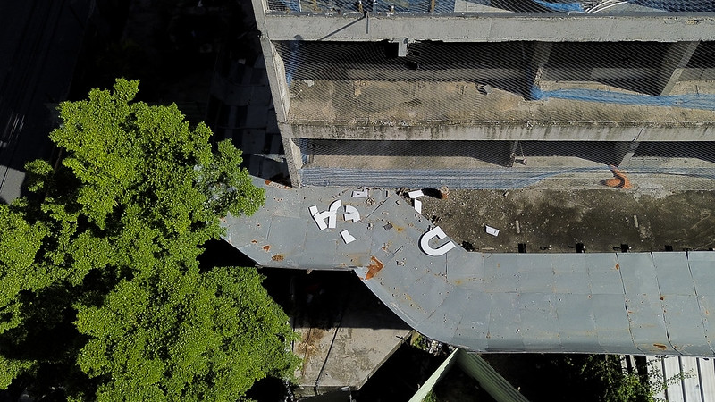 A imagem mostra uma vista aérea de parte de um prédio abandonado e degradado, com aparência de concreto cru e sem acabamento. O edifício tem pelo menos três andares visíveis, com redes de proteção penduradas nas estruturas abertas, sugerindo que o local já foi usado mas está atualmente desativado. Na parte inferior da imagem, há uma cobertura metálica curva, parcialmente enferrujada, sobre a qual estão espalhadas várias letras brancas grandes que caíram ou foram removidas de uma antiga sinalização. As letras parecem formar a palavra HAUT, embora estejam desordenadas. À esquerda, uma grande árvore verde preenche boa parte da imagem com sua copa densa, oferecendo um forte contraste com o cinza e a deterioração do edifício. A cena transmite abandono e descaso, sugerindo que o local já teve alguma importância ou uso anterior.