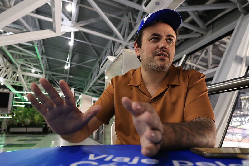 A imagem mostra o deputado federal Glauber Braga em um ambiente interno com estrutura metálica moderna e iluminação clara. Ele está sentado à frente de uma mesa azul, com a mão direita estendida e a esquerda parcialmente visível, como se estivesse gesticulando ou explicando algo. Glauber veste uma camisa polo marrom e um boné azul. Sua expressão facial é séria, mas calma, sugerindo que está engajado em uma conversa. À frente dele, sobre a mesa, há um celular. O cenário ao fundo indica que ele está em um local público, possivelmente o aeroporto dos Guararapes, devido à arquitetura e aos elementos metálicos visíveis no teto.