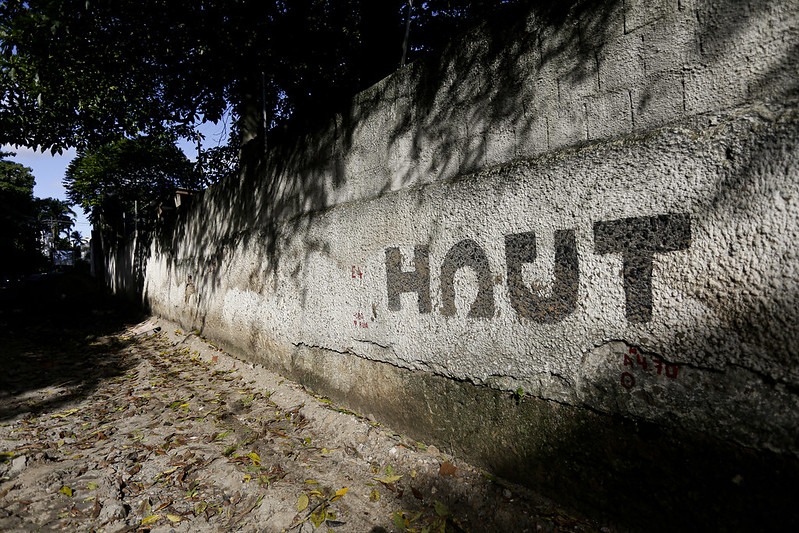 A foto mostra um muro de concreto alto e comprido, situado ao lado de uma rua de paralelepípedos com folhas secas espalhadas pelo chão. O muro, de cor clara e textura áspera, está parcialmente sombreado pelas copas de árvores que se estendem por cima dele, do lado de fora da imagem. No centro do muro, está pintada a palavra HAUT em letras grandes, escuras e em estilo simples, diretamente sobre a superfície áspera da parede. A iluminação do sol e as sombras das árvores criam um contraste marcante na cena, que transmite uma atmosfera silenciosa e um pouco desgastada. Ao fundo, do lado esquerdo da imagem, vê-se uma leve continuação da rua e vegetação ao longe.