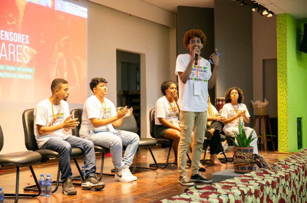 Na imagem, seis jovens participam de um evento em um palco. Todos vestem camisetas brancas com a frase “Jovens Defensores Populares”. Cinco estão sentados em fila, enquanto um jovem com cabelo crespo está em pé, falando ao microfone com um papel na mão. O grupo demonstra atenção e envolvimento; alguns batem palmas. Ao fundo, há uma tela de projeção com textos sobre direitos e defensores populares. O ambiente é colorido e iluminado, com destaque para uma parede verde e uma planta decorativa à frente do palco. Há garrafas de água no chão ao lado das cadeiras.
